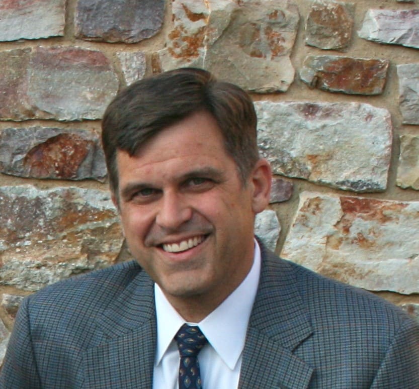 A man in a suit and tie smiles in front of a stone wall.